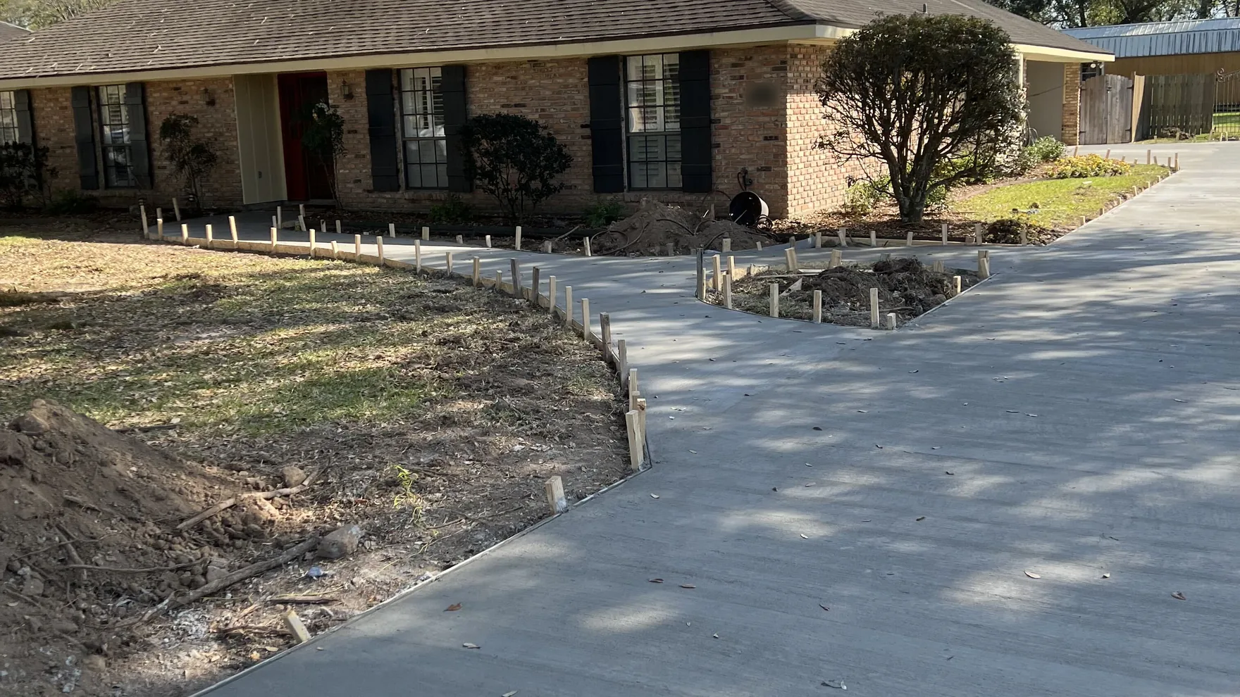 New concrete driveway and walk with form stakes in place in front of a one-story ranch house under oak limbs, concrete driveways