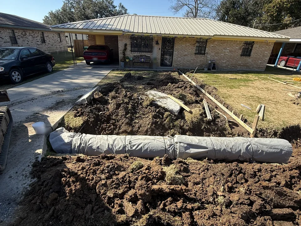 Driveway torn out to dirt in front of a brick house with a new fabric-wrapped culvert pipe laid across the front, concrete driveways