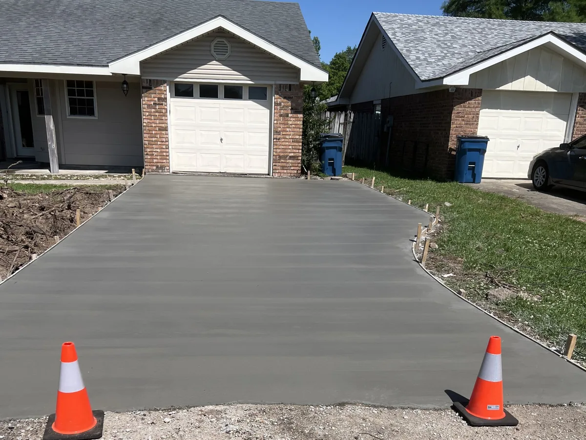 Freshly poured gray concrete driveway with a broom finish leading to a single garage, orange cones at the street end, concrete driveways