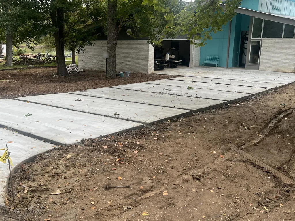 New concrete driveway with control joints sweeping up to a one-story house and carport under large oaks, concrete driveways