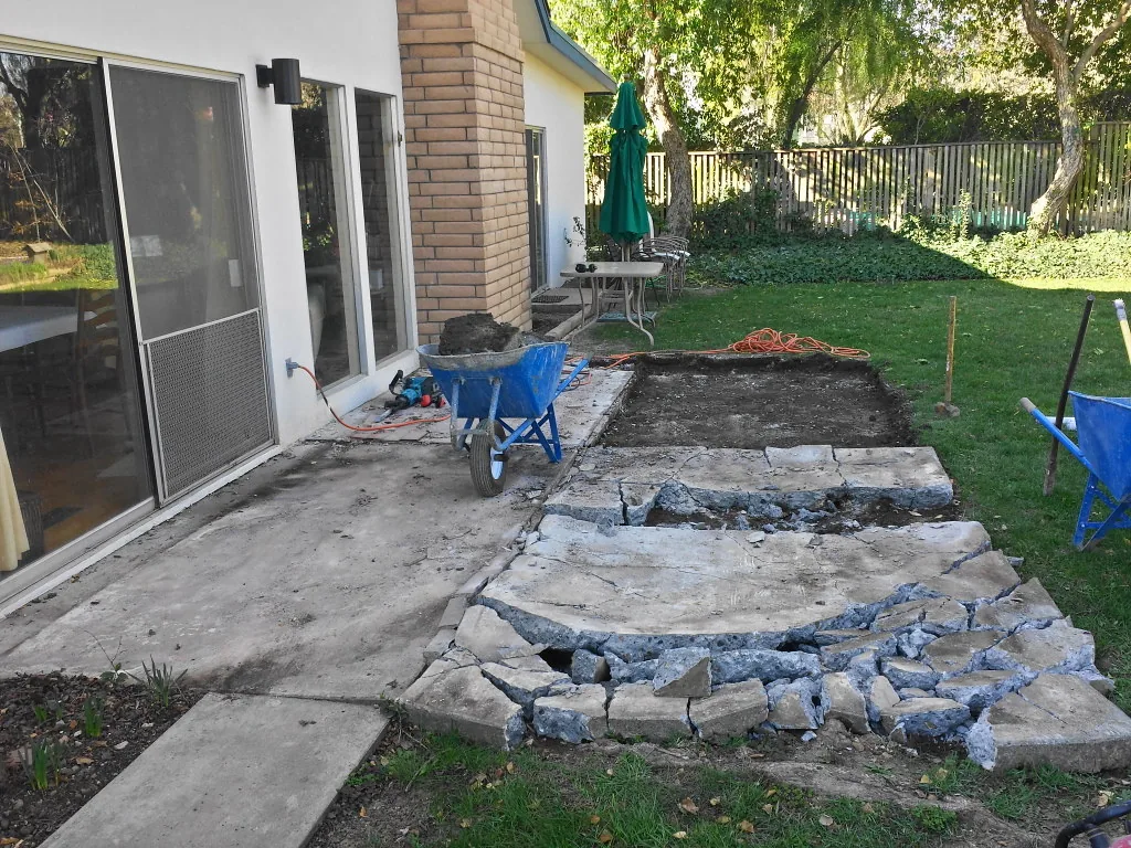 Backyard patio saw-cut into sections, with one section lifted out to bare dirt and a jackhammer and wheelbarrow on the slab, concrete removal and replacement