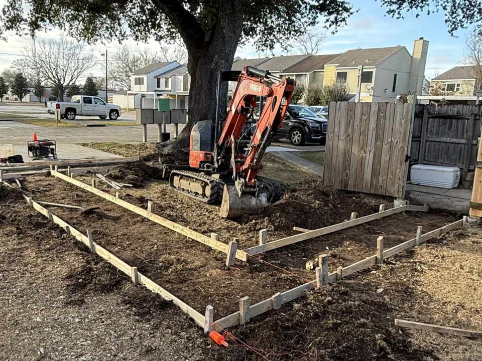 Mini excavator digging inside staked wood forms for a new slab in a backyard beside a wood fence, concrete slabs and pads