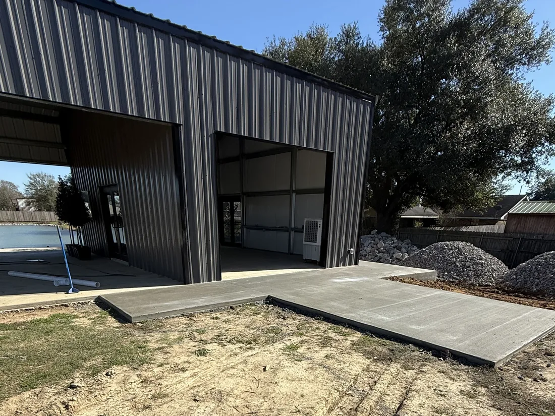 New concrete slab and apron poured at the open bay of a dark metal shop building under a live oak, concrete slabs and pads