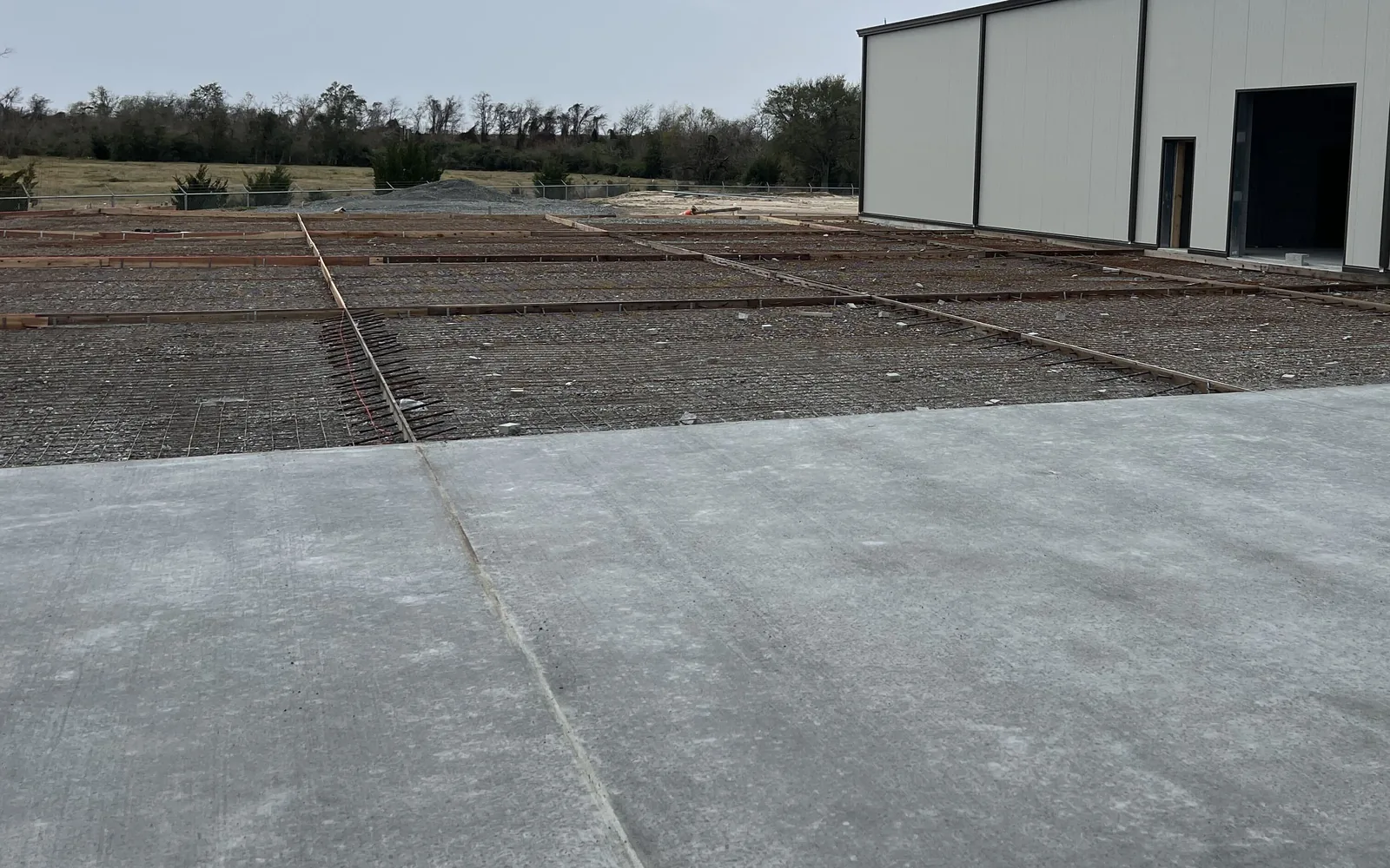 Slab poured in sections beside a metal building, with rebar mats set in the next bays between form boards, concrete slabs and pads