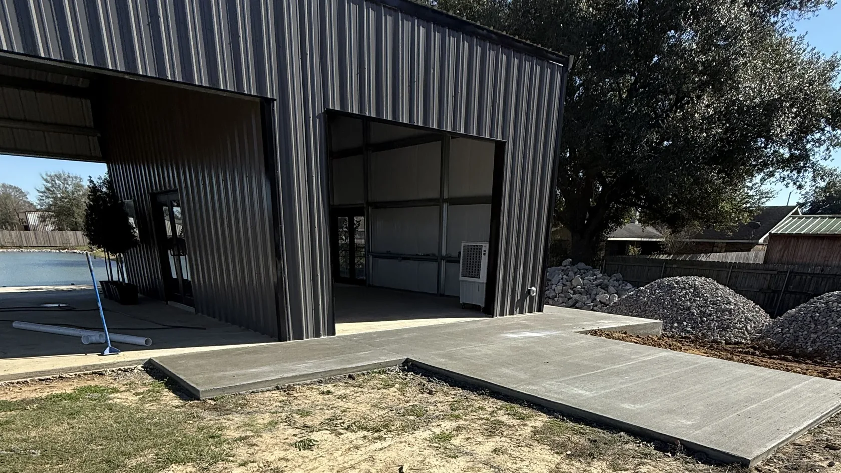 New concrete slab and apron at the open bays of a dark metal shop building with a live oak behind, concrete slabs and pads