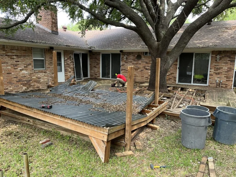 Old patio cover pulled down onto its deck frame beside a one-story brick house under a live oak, demolition