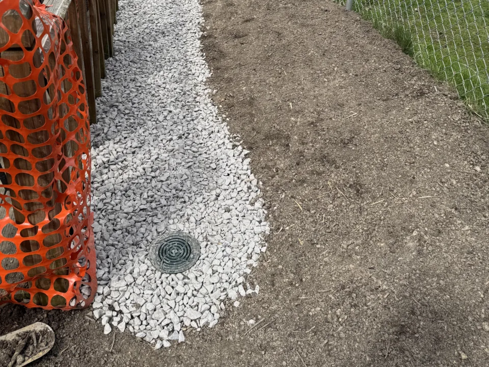 Finished French drain backfilled with white gravel along a wood fence, with a round drain grate at the end of the line, French drains