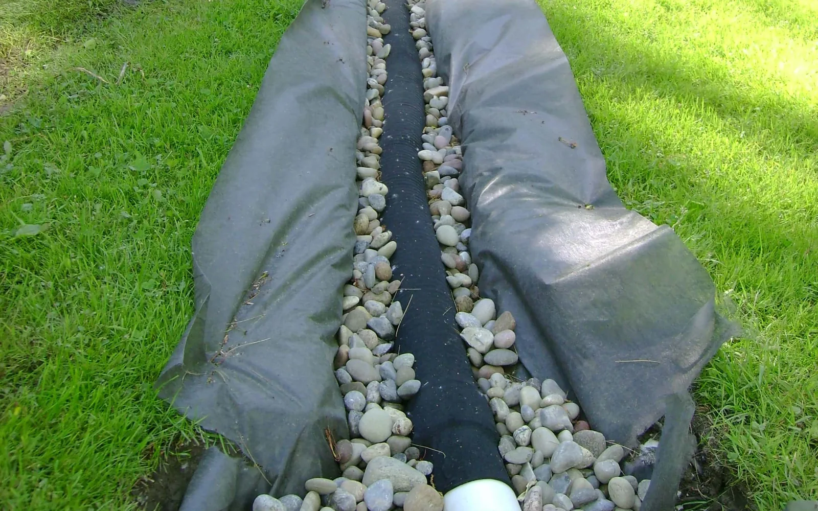 Drain trench lined with filter fabric, with washed rock placed over the pipe and a white pipe fitting, French drains