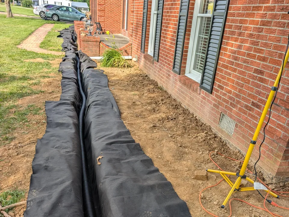Fabric-lined trench with black perforated drain pipe running along a one-story brick house, French drains