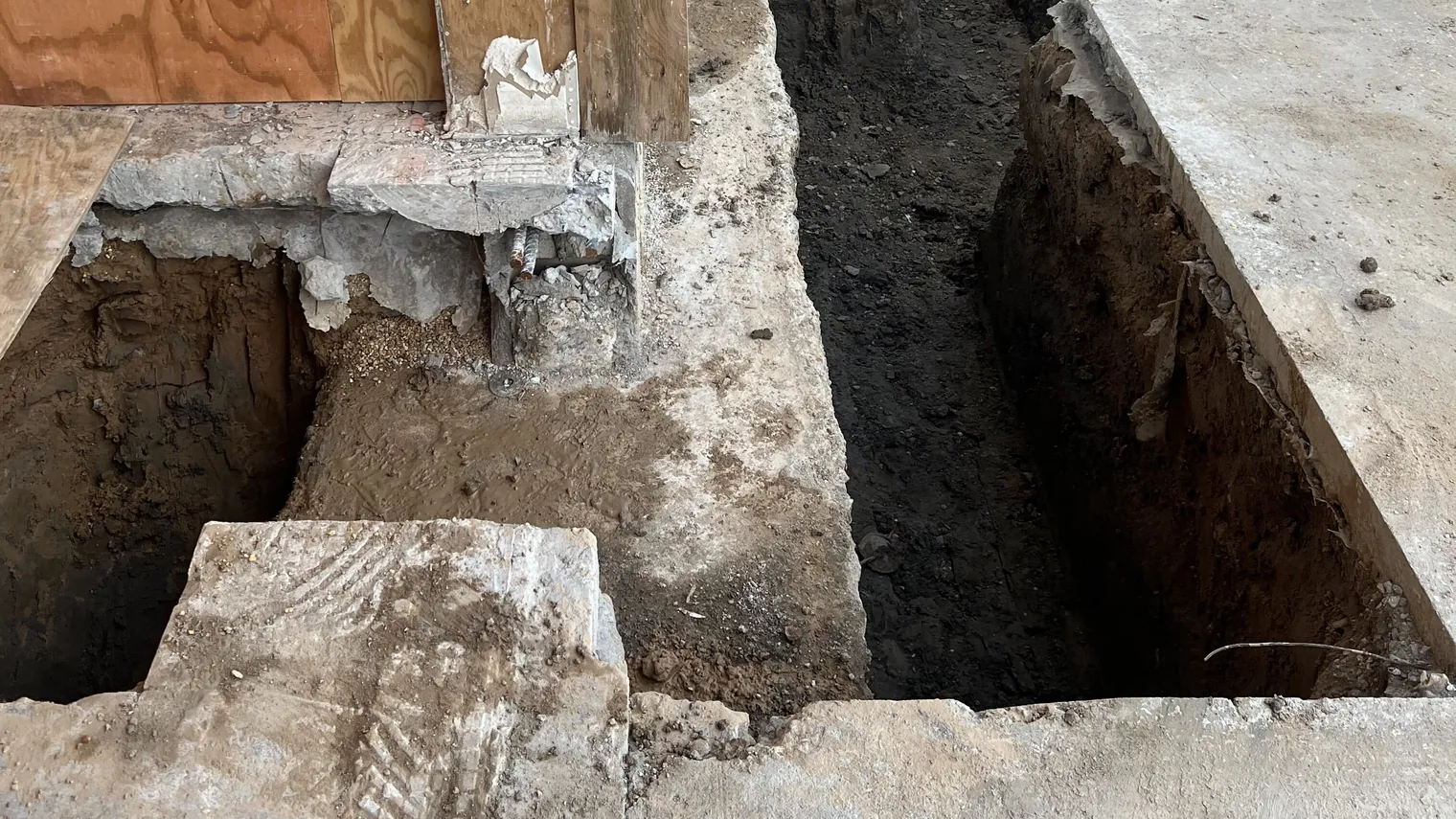 Footing trench cut through an old slab along a wall base, with plywood forms and broken slab edges, concrete foundations and footings
