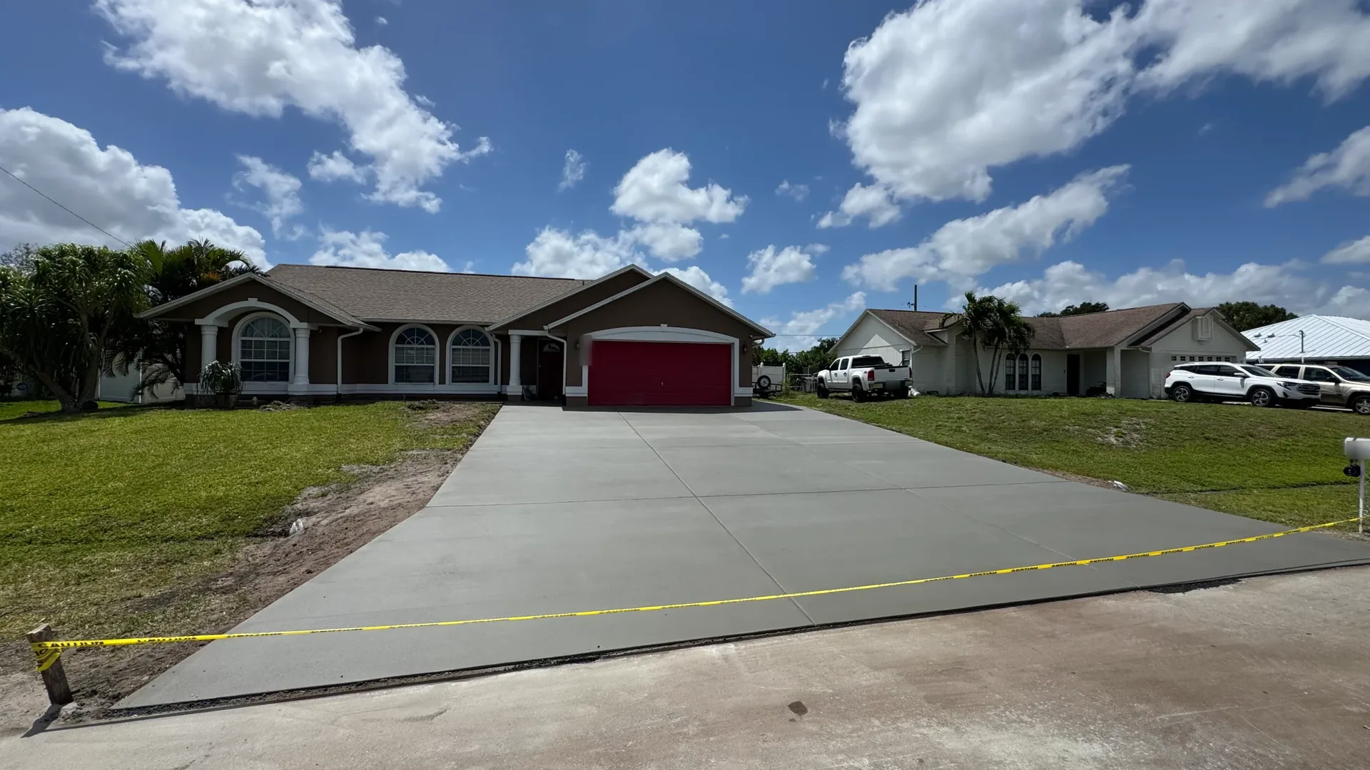 New concrete driveway with control joints and caution tape leading to a one-story house on a flat lot, concrete driveways