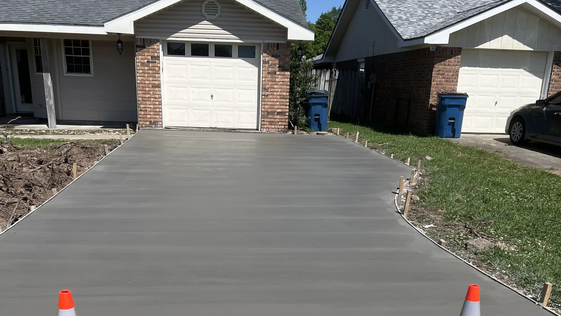 Freshly poured concrete driveway with a broom finish leading to a garage, orange cones at the street end, concrete driveways