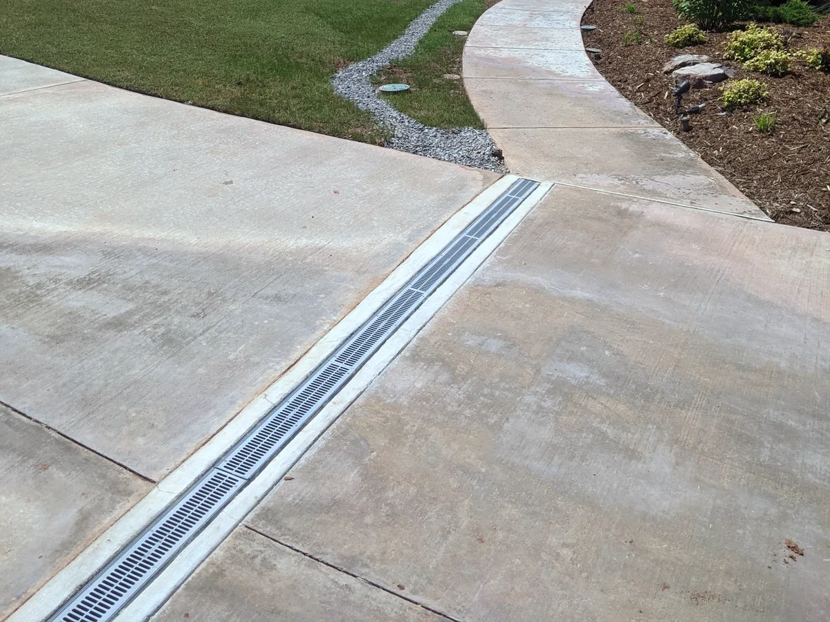 Channel drain with a gray grate set across a concrete driveway where it meets a curved walk, yard drainage