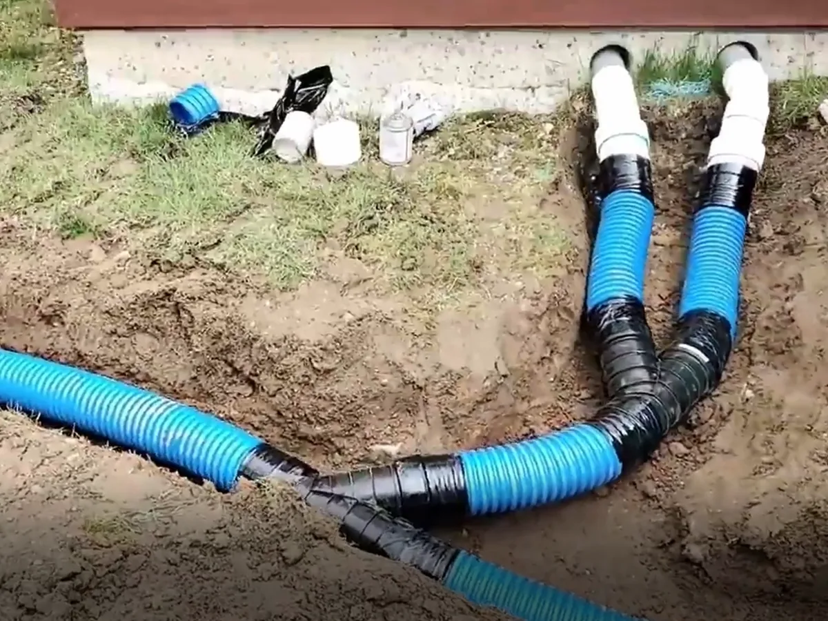 Two downspout lines joined into one corrugated drain line in a trench beside a house foundation, yard drainage